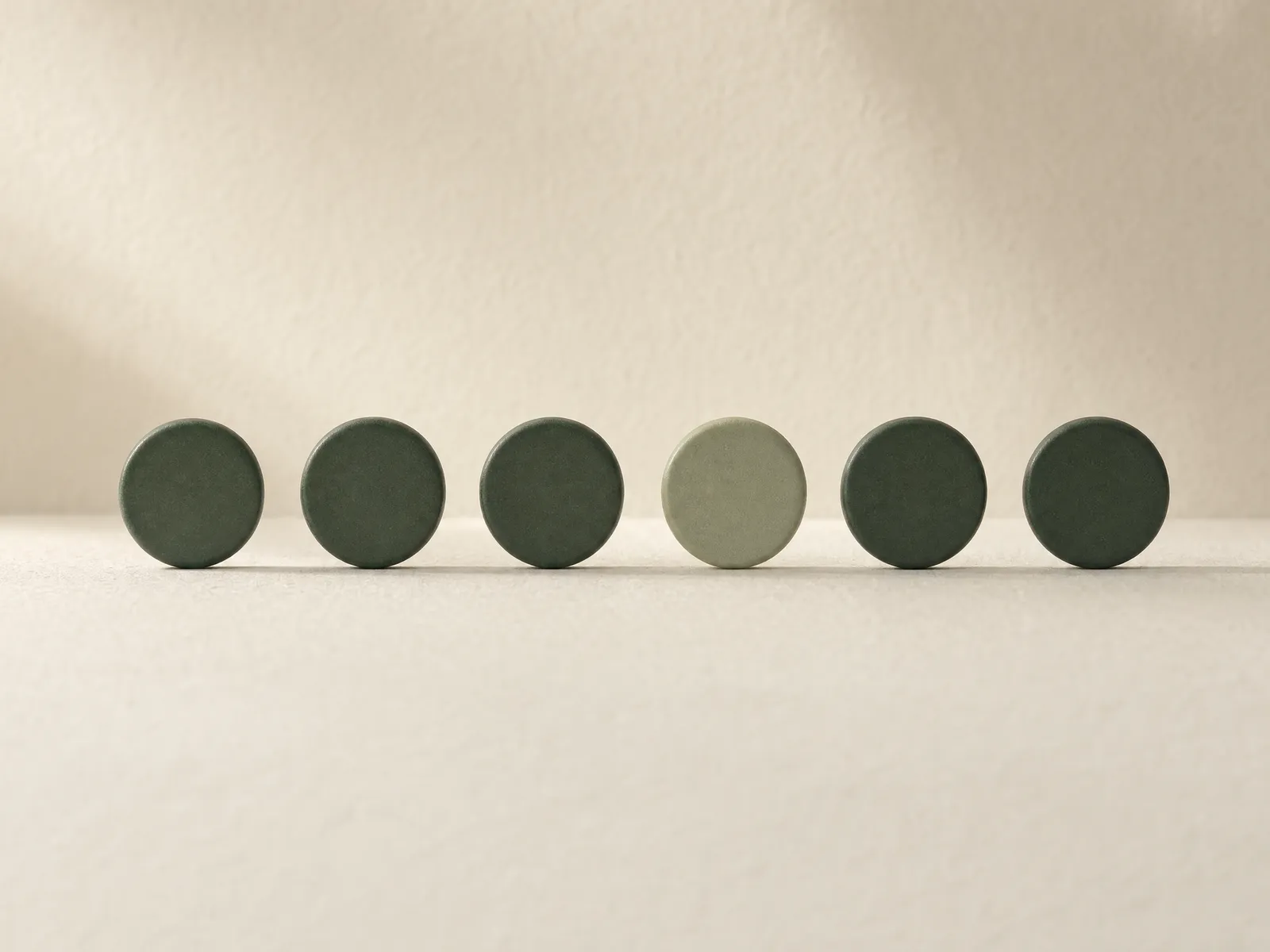 Six identical small ceramic tokens standing in a neat row on handmade paper, five deep green and one sage green.